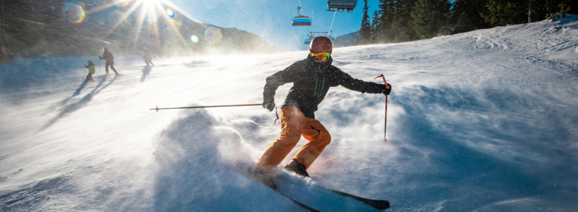 Jugendlicher Skifahrer bremst bei Wind auf einer sonnigen Piste mit sonnigen Berggipfeln und Gondelbahn im Hintergrund.