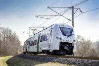 Sie sehen auf diesem Bild den Siemens Mireo Zug von vorn während einer Fahrt. Im Hintergrund sind nicht belaubte Bäume.