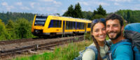 Ein Mann und eine Frau in Wanderkleidung stehen auf einer Wiese und lächeln in die Kamera.  Im Hintergrund sind Schienen zu sehen auf denen ein gelber Zug der Oberpfalzbahn fährt.