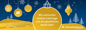 Auf dem Bild sieht man mehrere festlich dekorierte Weihnachtskugeln, die in einer weihnachtlichen Umgebung hängen. Eine der Kugeln trägt einen herzlichen Gruß, der allen Fahrgäste der oberpfalzbahn schöne Feiertage wünscht.