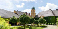 Das Bild zeigt das Schloss Reuthe im Sommer bei Sonnenschein. Im Vordergrund stehen Bäume in einer Parkanlage.