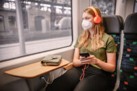 Eine Frau sitzt mit Maske im trilex. Währenddessen hört sie über Kopfhörer, die mit ihrem Handy verbunden sind Musik.