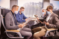 Vier Personen sitzen mit einem Mund-Nasen-Schutz im Zug. Sie beschäftigen sich mit Zeitung lesen, einem Tablet und einem Handy.