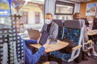 Es ist ein Zugabteil zu sehen, in dem mehrere Fahrgäste mit einem Mund-Nasen-Schutz sitzen und sich auf unterschiedliche Art und Weise Beschäftigen. Manche Lesen oder Arbeiten und nutzen dafür ein Tablet oder einen Laptop. Andere unterhalten sich.