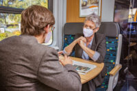 Zwei Fahrgäste sitzen sich in einem Vierer-Abteil gegenüber und tragen einen medizinischen Mund-Nasen-Schutz.
