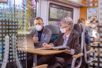 In einem trilex Abteil sitzen mehrere Fahrgäste, die eine Maske tragen. Zwei Fahrgäse sind im Fokus. Sie sitzen neben einander und unterhalten sich.