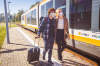 Zwei Fahrgäste mit Maske gehen auf dem Bahnsteig und ziehen einen Rollkoffer hinter sich her. Am Bahnsteig hält ein trilex Zug.
