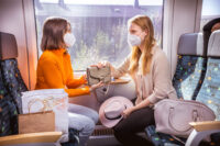 Zwei Frauen mit Maske sitzen sich im trilex gegenüber.