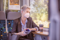 Frau sitzt mit Maske im Zug und schaut aus dem Fenster, vor ihr steht ein Kaffee aufm dem Tisch