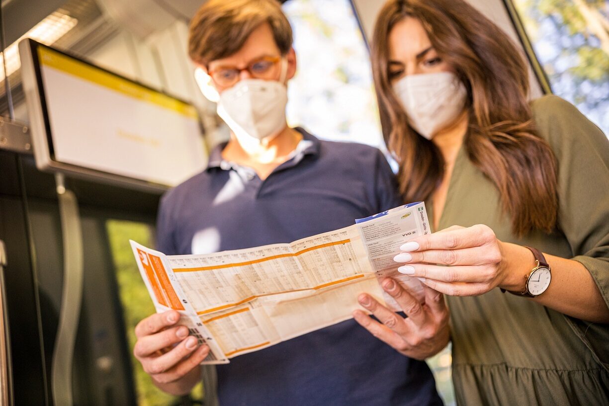 Paar trägt einen medizinischen Mund-Nasen-Schutz und liest einen Fahrplan im Zug.