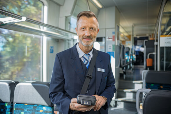 Ein Kundenbetreuer steht m Mittelgang des trilex und hält ein Fahrkartenverkaufsgerät in der Hand.