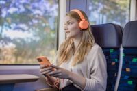Eine junge Frau sitzt mit Handy und Kopfhörern im trilex und hört Musik, während sie aus dem Fenster schaut.