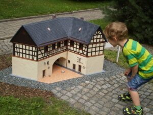 Klein Vogtland Das Bild zeigt ein Kind in einem grün-gelb gestreiften Shirt und schwarzen Shorts, das sich über ein detailliertes Miniaturmodell eines traditionellen Fachwerkhauses beugt. Das Modell befindet sich auf einer gepflasterten Fläche mit Gras und Bäumen im Hintergrund.