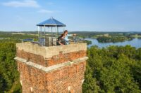 Das Bild zeigt einen Backstein-Aussichtsturm mit einem Metalldach und einem Geländer oben. Zwei Personen stehen auf der Aussichtsplattform und blicken auf eine malerische Landschaft mit dichten grünen Wäldern und einem großen Gewässer im Hintergrund. Der Himmel ist klar mit einigen verstreuten Wolken.