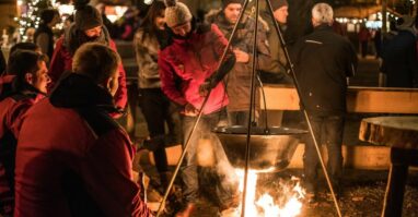 Christkindlmarkt Regen Menschen sitzen oder stehen um Lagerfeuer, über dem Glühwein erhitzt wird.