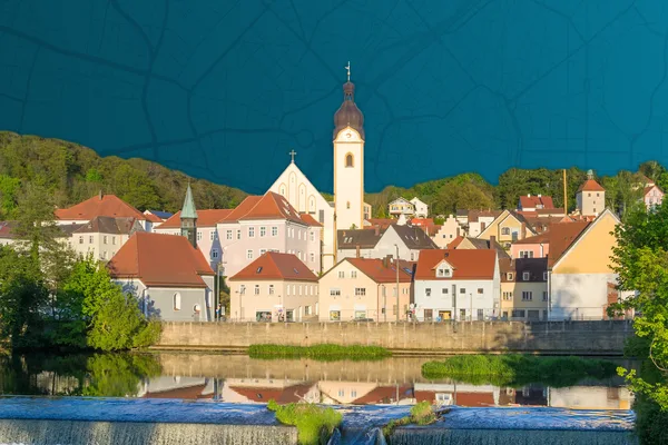 Fluss mit kleinem Wasserfall, dahinter Häuser mit roten Dächern und Kirchturm.