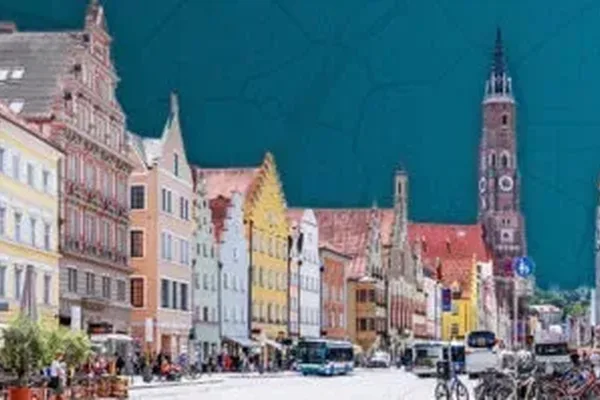 Das Bild zeigt eine belebte Straße mit bunten Häusern in Landshut. Im Hintergrund erhebt sich eine große Kirche mit hohem Turm.