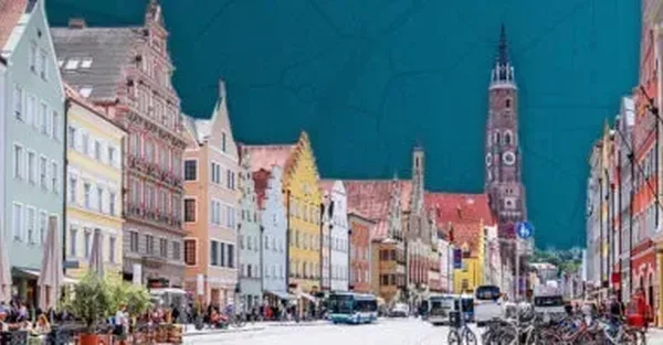 Obrázek zobrazuje rušnou ulici s barevnými domy v Landshutu. V pozadí se tyčí velký kostel s vysokou věží.