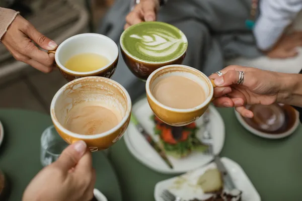 Vier Tassen mit Kaffee, Matcha und Tee werden über einem Tisch zusammengestoßen, Teller mit Kuchen im Hintergrund.