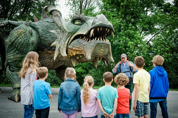 Große Drachenfigur mit geöffnetem Maul und Zähnen, umgeben von grünen Bäumen, Besuchergruppe davor.