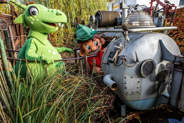 Zwei Figuren in Kostümen, darunter ein grüner Drache und ein Charakter mit roter Mütze, neben einer alten Metallmaschine im Freien.