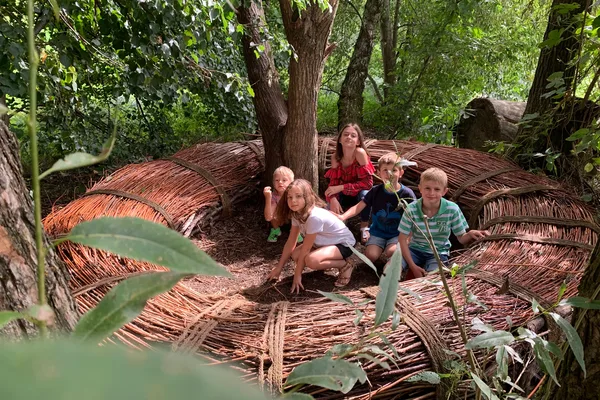 Kinder sitzen in einem großen, kreisförmigen Nest aus geflochtenen Zweigen unter Bäumen im Wald.