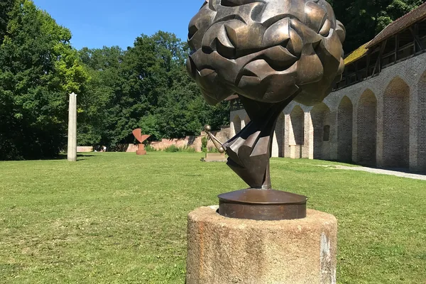Bronzeskulptur auf Sockel im grünen Garten des KOENIGmuseums, historische Mauern im Hintergrund.