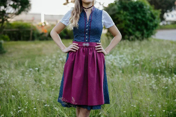 Person trägt ein traditionelles Dirndl in Blau und Rot, stehend auf einer grünen Wiese.