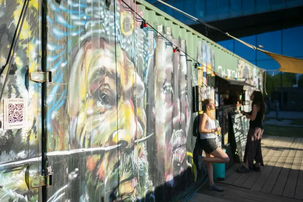 Container mit farbenfrohem Graffiti vor einem improvisierten Outdoor-Bereich mit Holzplanken und Sonnensegeln. Im Vordergrund sind zwei Personen im Gespräch.