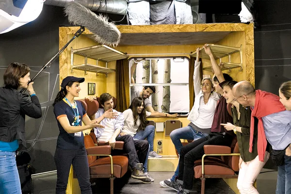 Filmset in der Bavaria Filmstadt mit nachgebautem Zugabteil, roten Sitzen und Gepäckablagen, Tonangel und Beleuchtung im Einsatz.