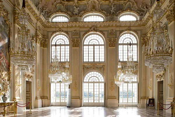 Großer barocker Saal mit hohen Fenstern, goldenen Verzierungen und mehreren Kristalllüstern.