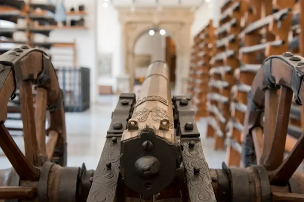 Nahaufnahme einer verzierten Kanone auf Holzrädern in einer Ausstellungshalle mit Waffenregalen.
