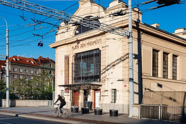 Historisches Bahnhofsgebäude mit der Aufschrift ‚Plzeň Jižní Předměstí‘ und Oberleitungen, davor ein Radfahrer.