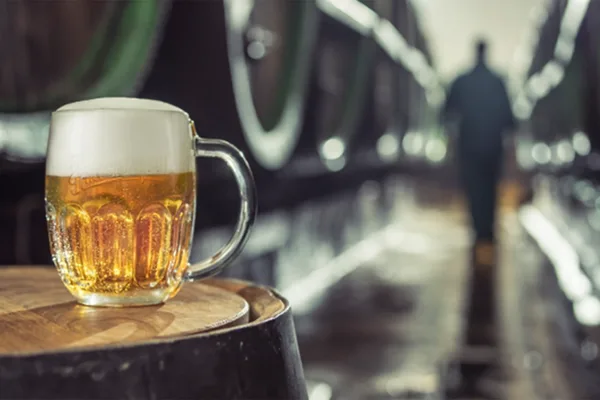 Bierkrug mit Schaum auf einem Holzfass, im Hintergrund Brauereifässer und unscharfe Person.