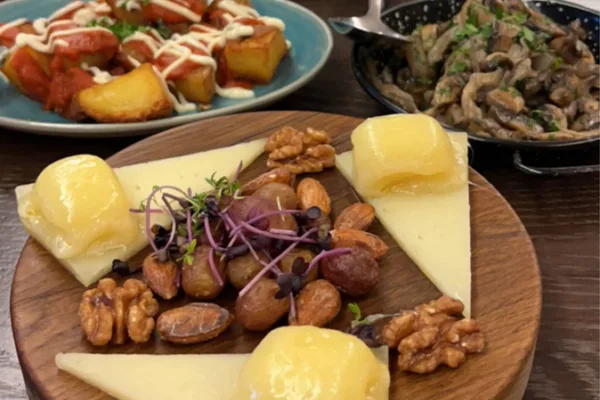 Holzteller mit Käse, Nüssen und Trauben, im Hintergrund weitere Tapas-Gerichte.