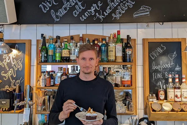 Person hält ein Stück Kuchen vor einer Bar mit Spirituosen und Tafeln mit handgeschriebener Speisekarte.