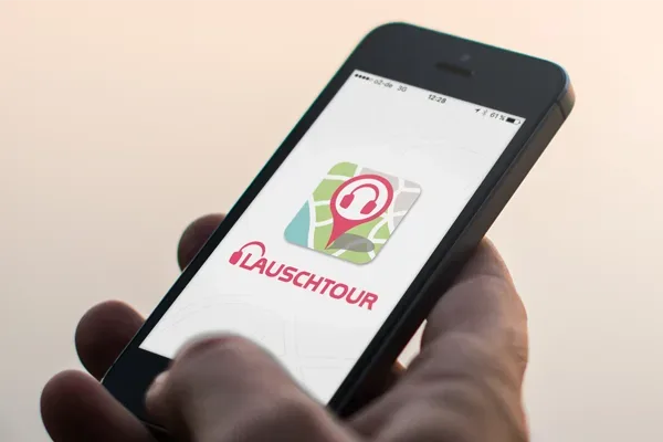 Hand hält ein Smartphone mit geöffneter Lauschtour App, angezeigt wird das App-Logo einer Audioguide-Anwendung mit Kartenmotiv und Kopfhörersymbol.