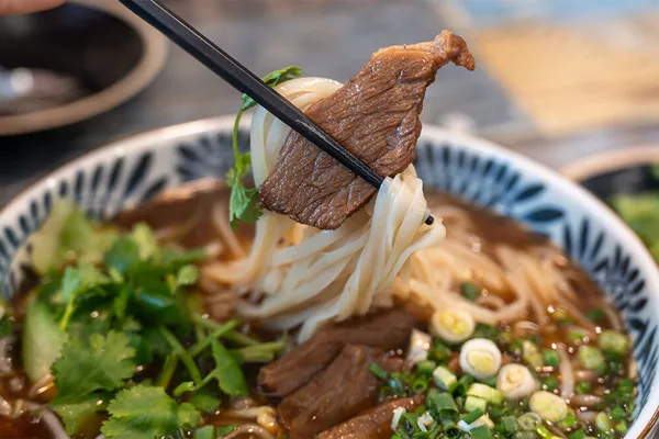 Schüssel mit Nudelsuppe, Rindfleisch und frischen Kräutern, Essstäbchen heben ein Stück Fleisch.