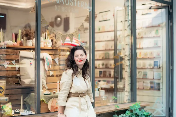 Außenansicht eines liebevoll dekorierten Ladens mit Holzregalen, Karten und Geschenkartikeln. Im Vordergrund ist die Besitzerin des Concept Stores zu sehen.