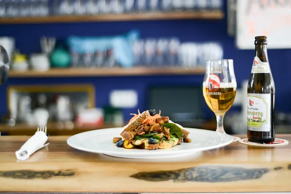 Teller mit kreativ angerichtetem Gericht auf einer Holztheke, daneben ein Glas Bier und eine Flasche Naabecker.