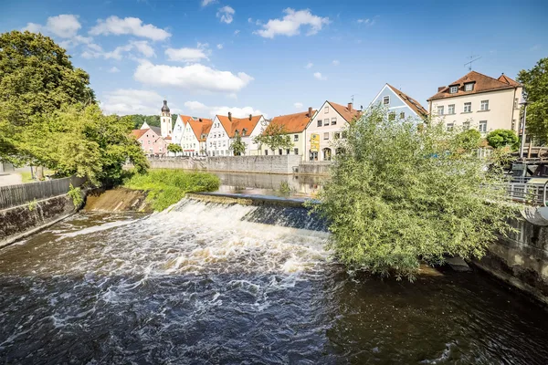 Blick auf Fluss mit kleinem Wasserfall, umgeben von Bäumen und Häusern mit roten Dächern im Hintergrund.