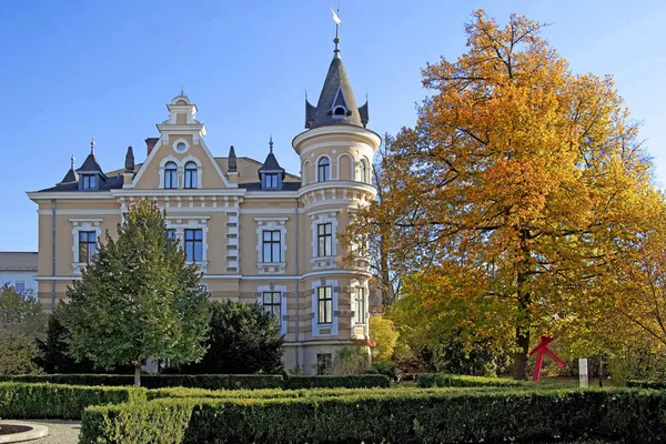 Prächtige Villa mit Türmchen und gelber Fassade, umgeben von gepflegtem Garten und herbstlichem Baum.