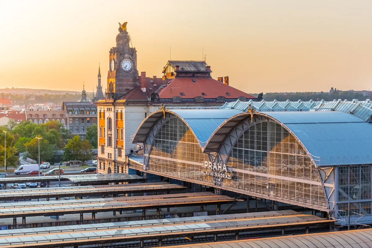 Glasdach des Bahnhofs mit Uhrturm und roten Dächern, dahinter die Stadt Prag im warmen Licht.