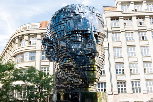 Spiegelnde Skulptur des Franz-Kafka-Kopfes aus Metallplatten vor modernen Gebäuden.
