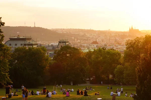 Menschen sitzen auf einer grünen Wiese im Park, im Hintergrund die Prager Skyline bei Sonnenuntergang.