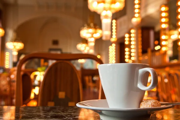 Weiße Kaffeetasse auf einem Marmortisch in einem eleganten Café mit warm beleuchteten Kronleuchtern im Hintergrund.