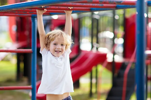 Kind hängt an einer roten Kletterstange auf einem Spielplatz mit weiteren Spielgeräten im Hintergrund.