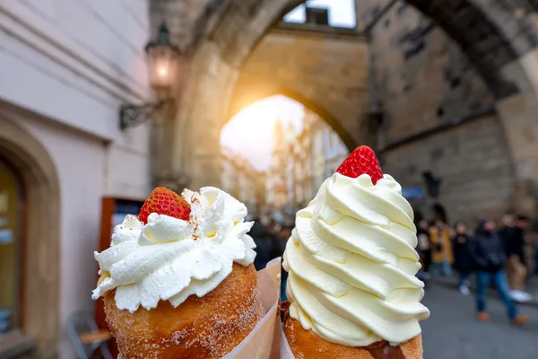 Zwei Trdelník-Gebäcke mit Sahne und Erdbeeren vor einem historischen Torbogen in der Prager Altstadt.