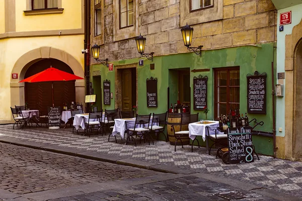 Grüne Hausfassade mit Tischen, Stühlen und Speisetafeln auf Kopfsteinpflasterstraße.