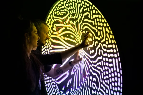 Person berührt eine leuchtende, farbenfrohe Installation mit organischen Mustern.